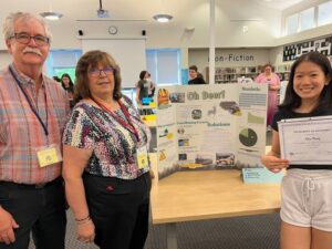 A grey-haired man and a woman stand next to a table that has a poster with the title "Oh Deer!" A girl stands on the other side of the table holding a certificate from the League of Women Voters that says "Excellence in Advocacy awarded to Elsa Meng.