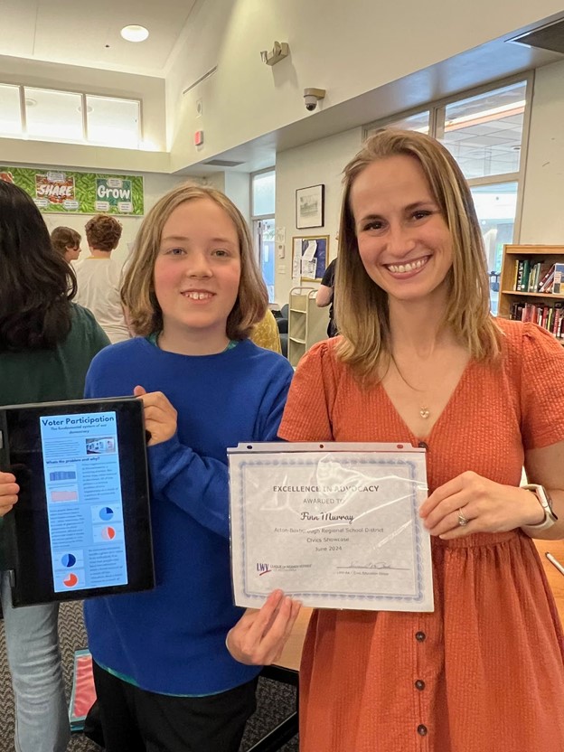 A young person holds a notebook that describes their project (Voter participation) stands next to a woman who is holding a certificate from the League of Women Voters that says "Excellence in Advocacy awarded to Finn Murray.