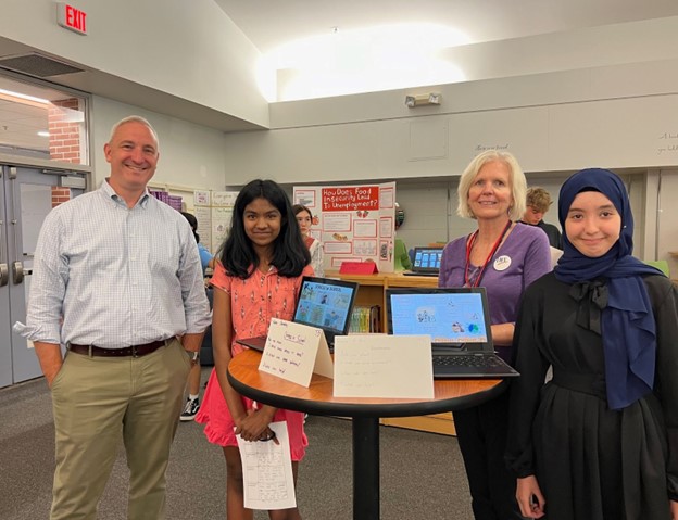 A grey-haired man wearing a sportshirt and slacks stands next to a smiling girl with long black hair, a grey-haired woman, and a smiling girl wearing a hijab. They are standing around a high table with two computers and signs that describe their projects.