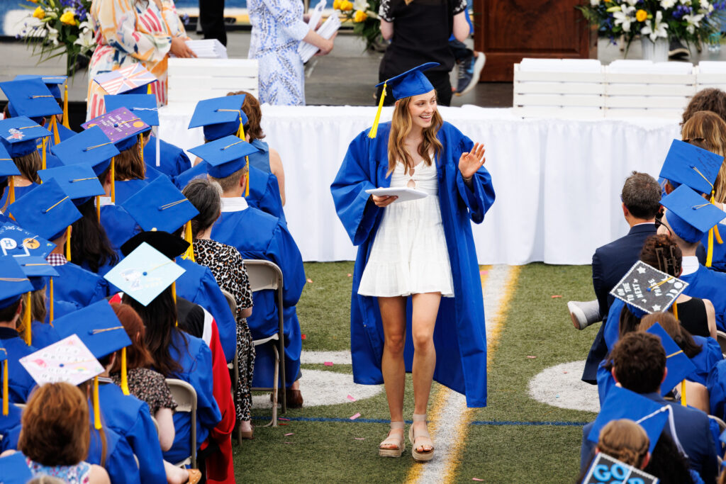 A young woman walks down the aisle sporting a blue graduation robe and cap. She is holding her diploma in one hand and waving at the seated graduates as she heads back to her seat.