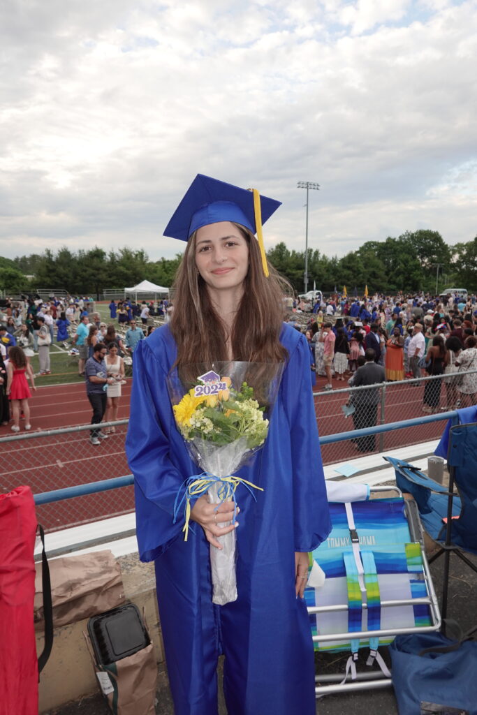 A young woman stands on Leary Field holding a bouquet of flowers and wearing her blue graduation cap and gown.