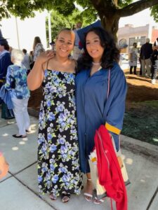 Two women pose proudly. The mom is smiling broadly and the daughter has her arm over her mother's shoulders. She's wearing the Minuteman blue and gold cap and gown.