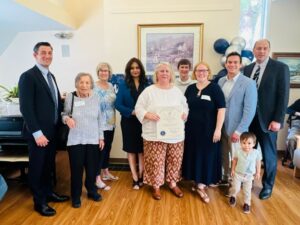 A group of people stand in a room with blue and silver balloons in the background. A woman stands in the middle of the group holding a large certificate from the Commonwealth of Massachusetts. A small boy is beginning to leave the crowd and walk towards the photographer.