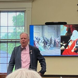 A man wearing a pink shirt and a bow tie speaks to an audience. There is a slide up on the screen behind him.