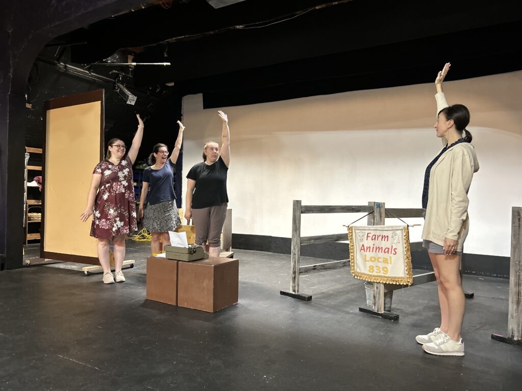 On a stage, three women are standing around a box where a typewriter sits. Another woman is on the other side of the stage and all of them have their hands raised. A sign hanging on a fence post reads "Farm Animals Local 839."