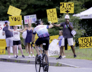 A rider with a Pan-Mass Challenge jersey is waving to people on the side of the side of the road waving brightly colored inspirational signs such as You Rock!,and U Ride, We Live.