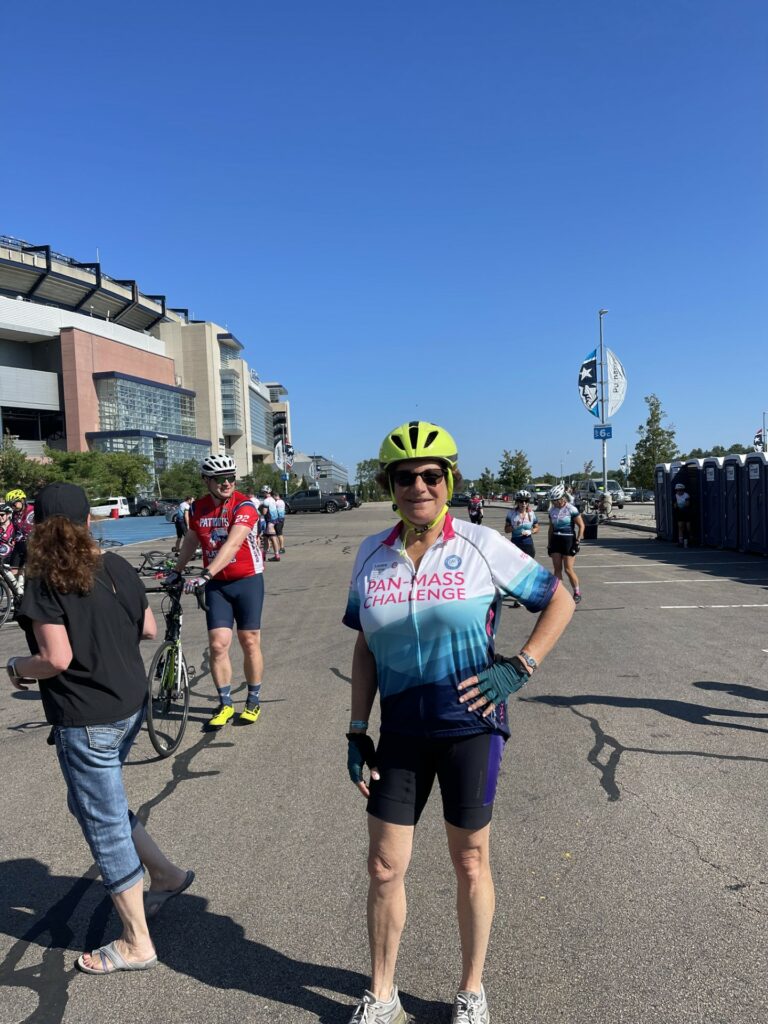 A woman in a pink and blue PMC jersey stands in froont of Patriot Place. There are other riders in the background.