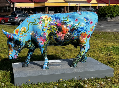 A richly decorated cow sculpture. The cow is blow and painted with flowers and other decorative elements. A grocery store is in the background.