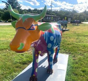 A cheerily painted cow statue -- this one has a mostly orange face, turquoise horns, and a purple body with blue spots. There are also some other cow statues in the background.