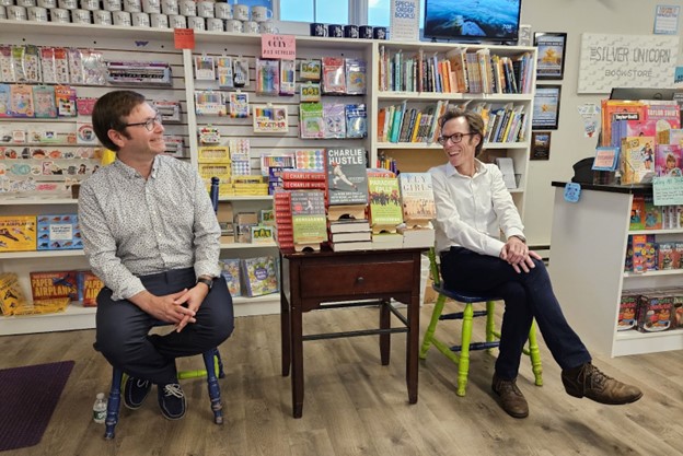 Two men wearing dress shirts are sitting in front of book shelves at Silver Unicorn Bookstore. Piles of books, all written by either Alex Speier or Keith O'Brien, are stacked on a table between them.