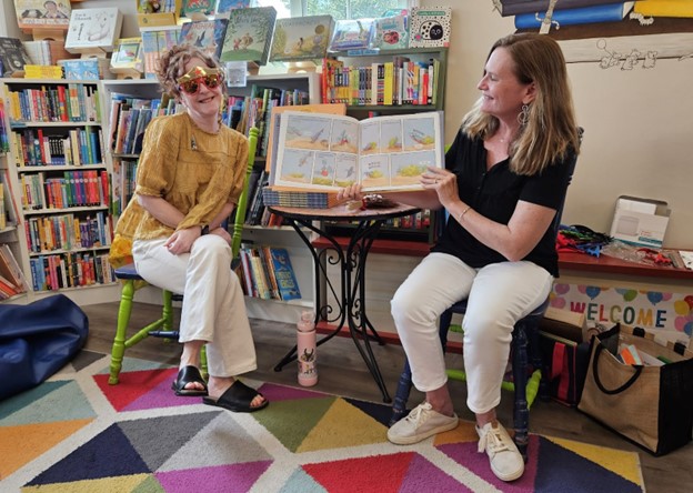 Two woment read a picture book in the Children's section of Silver Unicorn bookstore. One of the women is wearing red sunglasses with a crown on top.