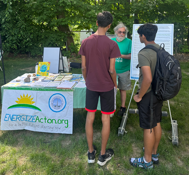 A man wearing a green t-shirt is speaking with two young men in front of the EnergizeActon booth.