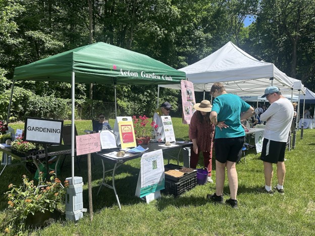 A booth at an outdoor fair with a green awning. A woman with a straw hat is showing something to a couple of fair-goers.