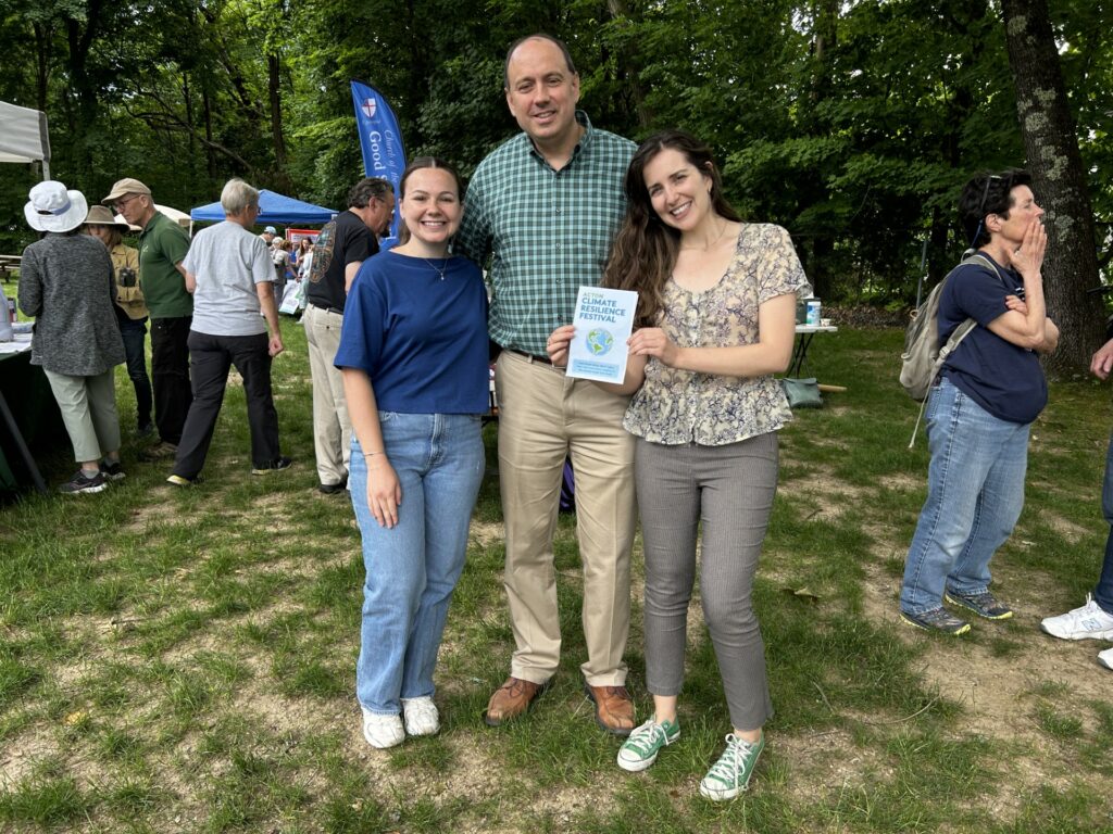 A tall man and two much shorter women pose. One of the women holds a Climate Resilience Festival poster.