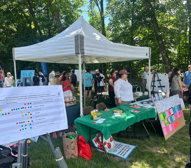 A booth at an outdoor fair with a poster on the left and a table covered with a green cloth. A woman stands behind the table.