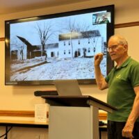 A man wearing a green polo shirt stands in front of a screen at the Acton Memorial Library. The screen shows a picture of the Edwards house from many years ago.