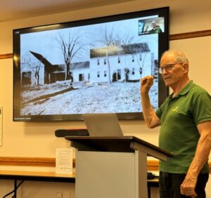 A man wearing a green polo shirt stands in front of a screen at the Acton Memorial Library. The screen shows a picture of the Edwards house from many years ago.