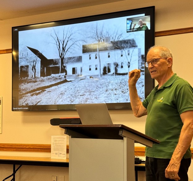 A man wearing a green polo shirt stands in front of a screen at the Acton Memorial Library. The screen shows a picture of the Edwards house from many years ago.