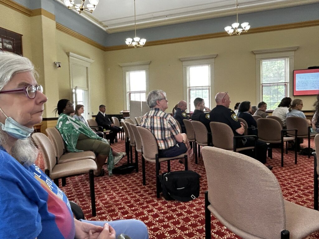 A number of people sitting facing the front of Room 204. The room is fairly full and you can see Police Chief James Cogan, Council on Disabilities member Lisa Franklin, and Select Board Member David Martin in the audience.