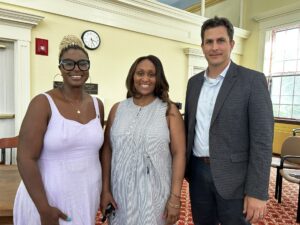 Two smiling women and a man pose in Room 204 of Town Hall. The women are wearing summer dresses, and the man (Town Manager John Mangiaratti) is wearing a sport coat. He looks very warm.