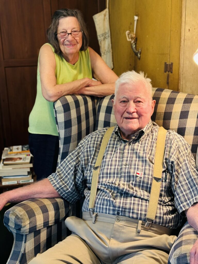 Two older people pose on a plaid armchair. He is sitting, she is resting her arms on the top of the chair.