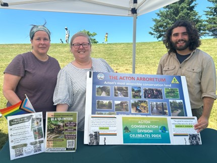 Two women and a man stand under a booth in front of a large Acton Arboretum poster.