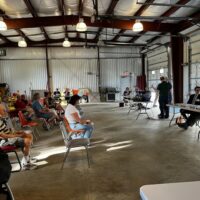 A cavernouse metal shed (the Acton Water District headquarters) where the audience is seated on the left, and various town officials (Finance Committee, Water District Commission) are seated at the tables on the right.