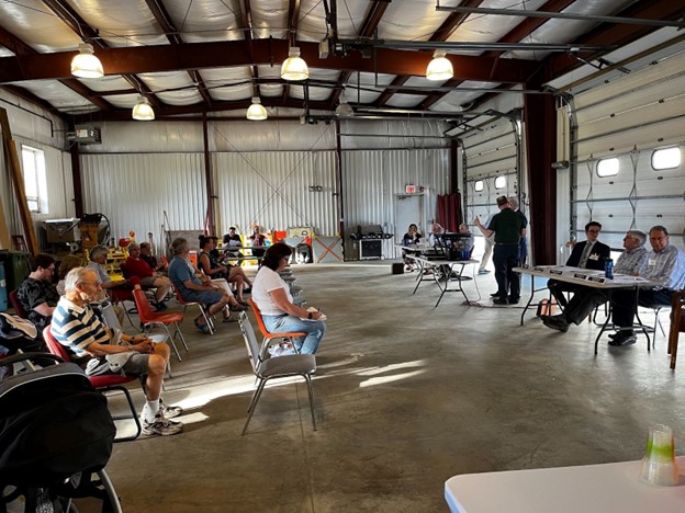 A cavernouse metal shed (the Acton Water District headquarters) where the audience is seated on the left, and various town officials (Finance Committee, Water District Commission) are seated at the tables on the right.