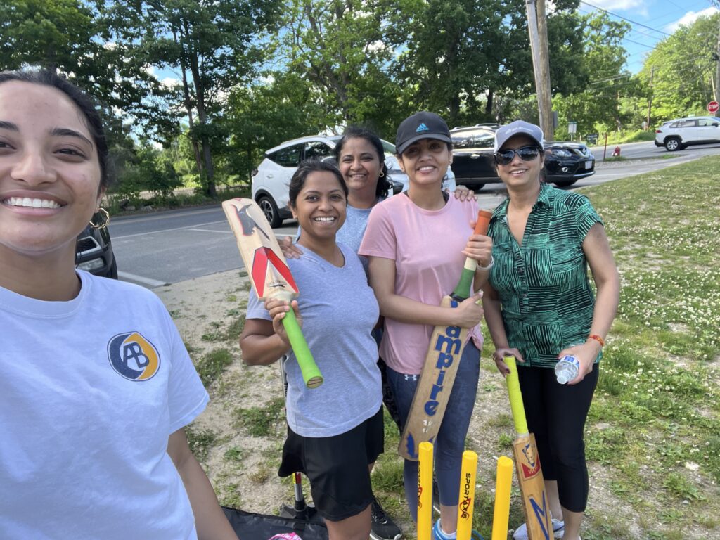 A group of smiling women with cricket bats in front of a cricket wicket.