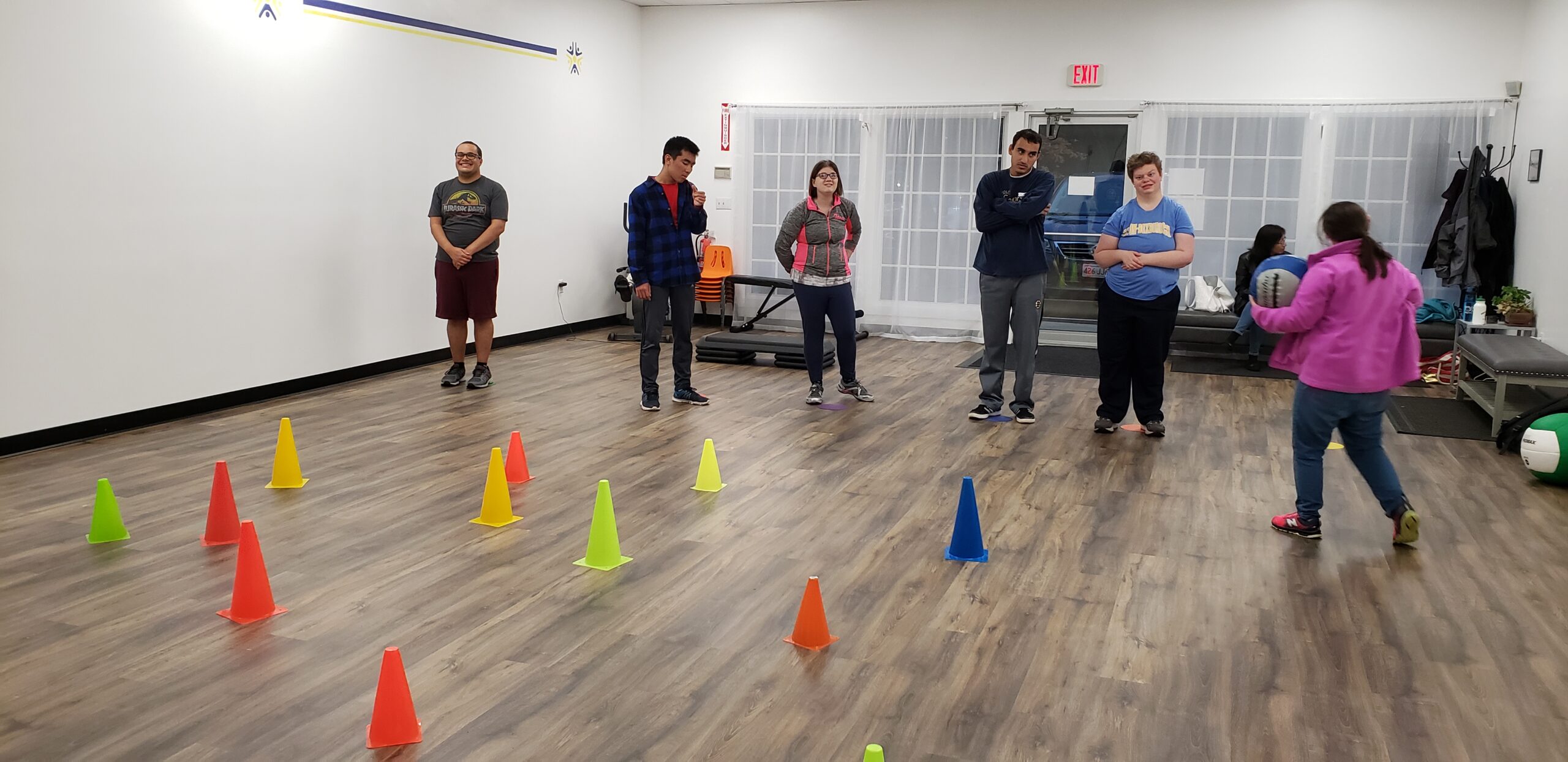 A group of people are standing in a gym. There are colored cones on the floor and a woman in a pink jacket brings a ball over to the clients.