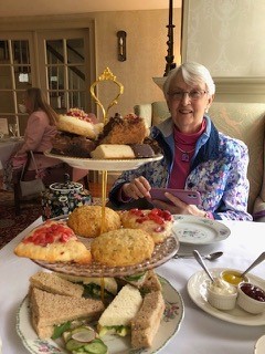 A woman sits behind a three-tiered china cake stand that is piled high with scones, sandwiches, and other delicacies that befit a proper English tea.