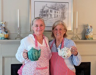 Two smiling women are wearing aprons and holding pretty teapots. They are standing in front of a white mantel decorated with pottery and candlesticks.