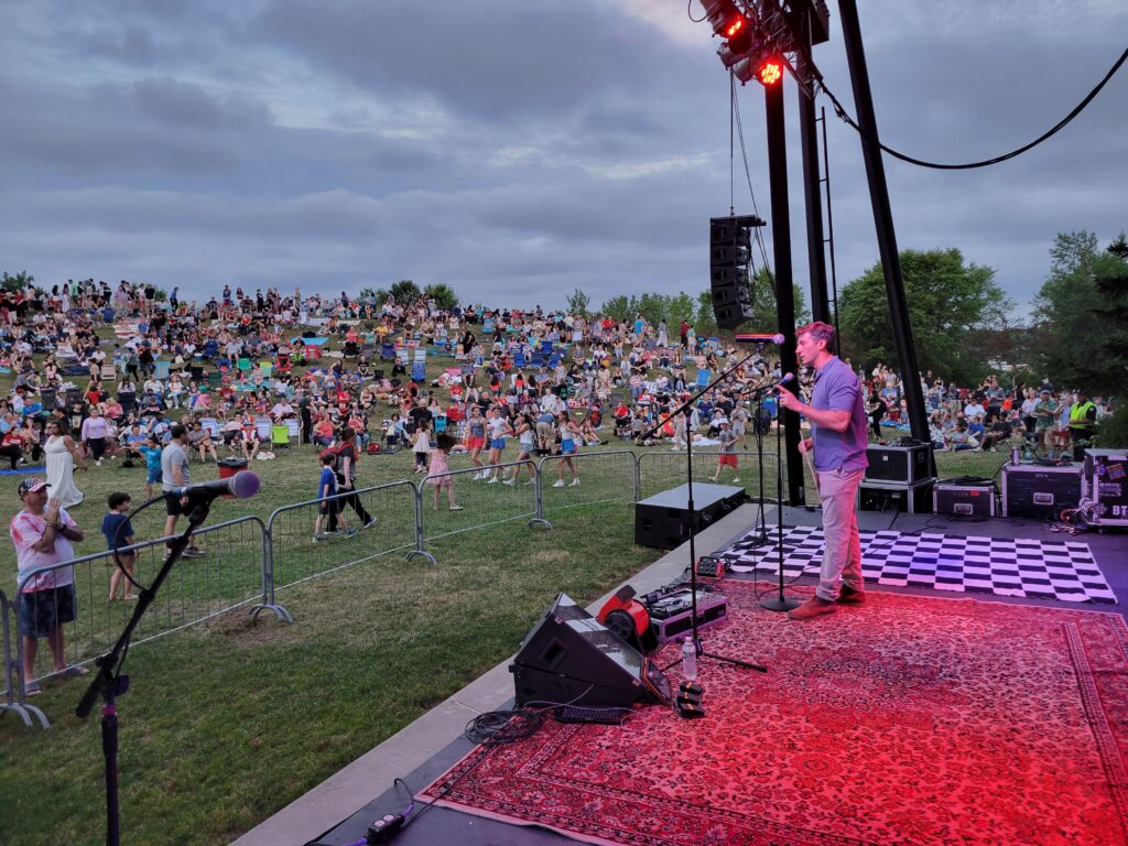 A man stands at a mic in the amphitheater. In the background, the hill is covered with people waiting for the band and fireworks.