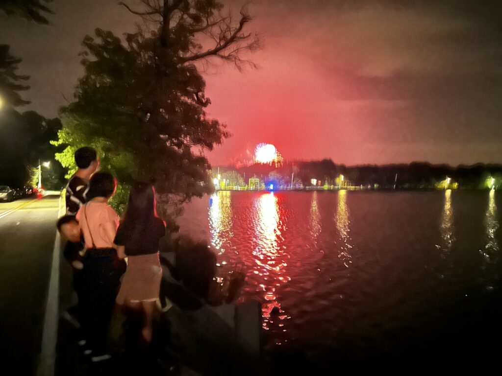 The sky and Nagog Pond glow red from fireworks at NARA. A family stands on the edge of the road to watch.