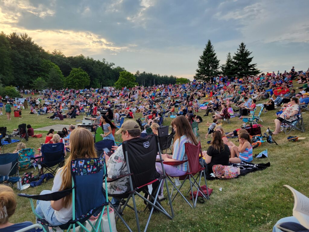 The hill at NARA park amphitheater is coverd with people sitting on blankets, sitting in chairs, and walking around. The sky is cloudy, but clearing.