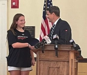 A man wearing a suit and tie stands at a podium. He is presenting an award to a young woman dressed in a t-shirt and sneakers.