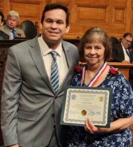 A smiling man and woman stand together. The woman is proudly holding a certificate.