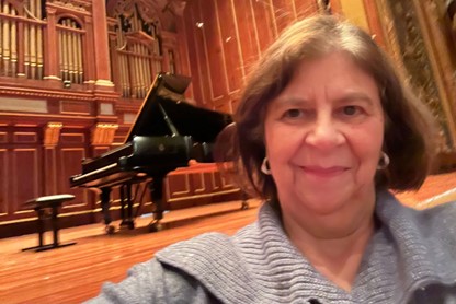 A smiling woman stands in front of a piano. The piano is up on stage in a concert hall.