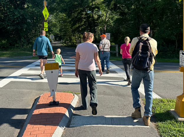 A group of people from the back, both adults and kids, cross a sidewalk on a sunny day.