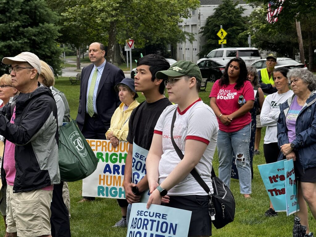 A group of people stand on a lawn all looking in the same general direction. Some people carry signs supporting reproductive rights.