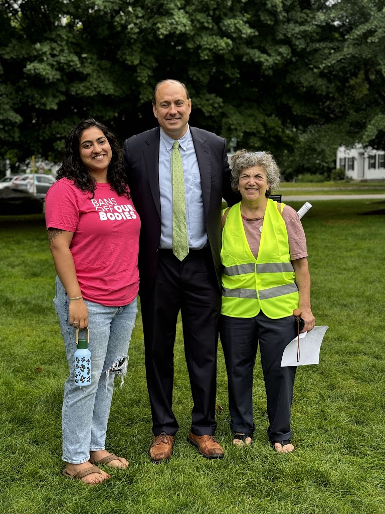 A man stands with two women on a lawn. One women wears the bright yellow vest of a rally marshall.