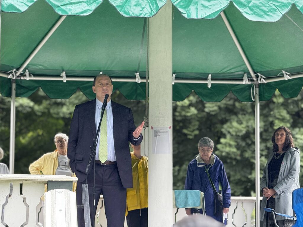 A man wearing a suit and tie speaks into a mic underneath a tent. A group of women are listening in the background.