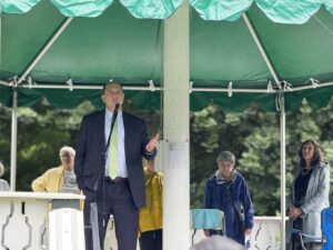 A man wearing a suit and tie speaks into a mic underneath a tent. A group of women are listening in the background.