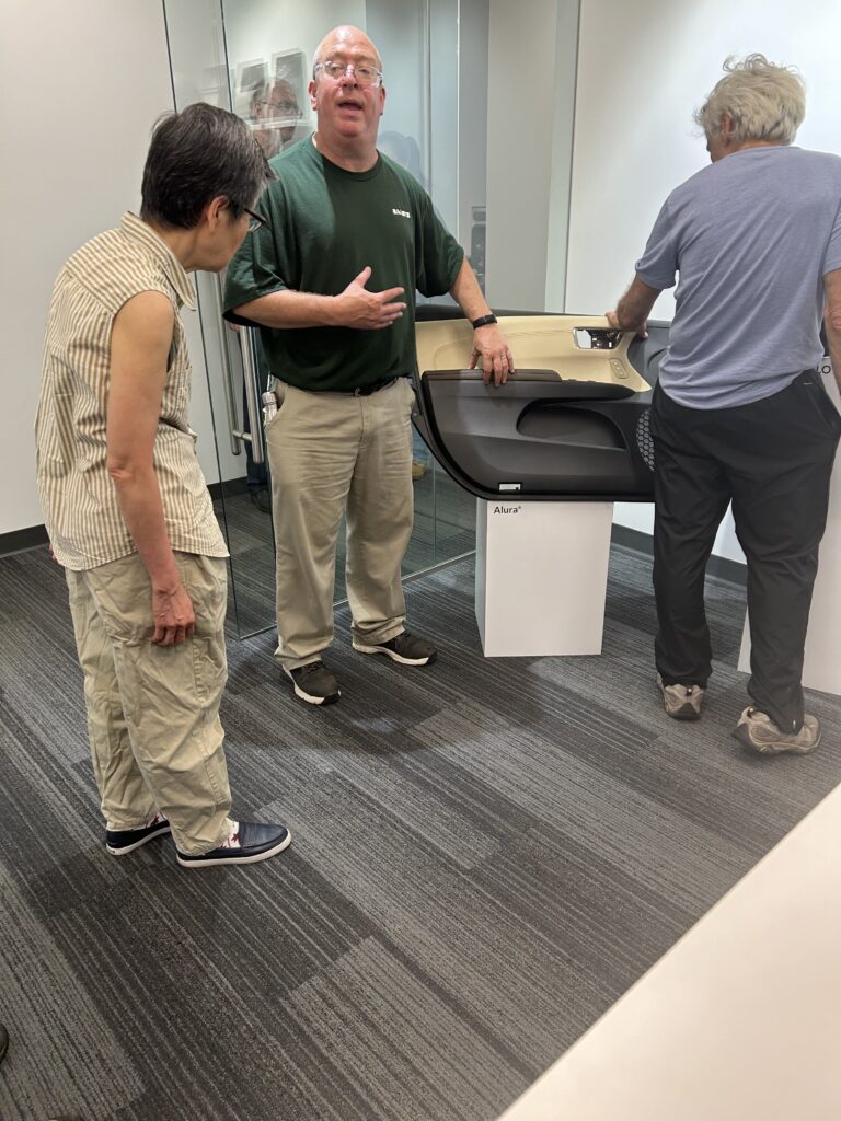 A man in a green t-shirt shows off a car door covered with Haartz fabric. The door is on a stand labeled Alura (one of the Haartz products) and two people are examining the fabric on the door.