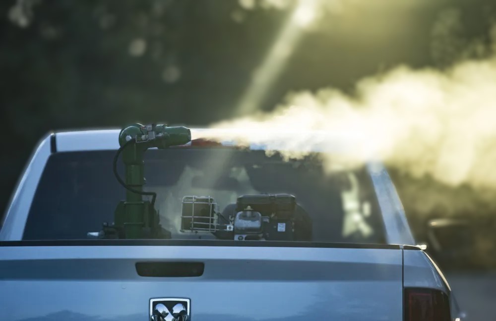 A white truck from the back with a fogging machine in the truck bed. The fogging machine of shooting out mosquito spray.