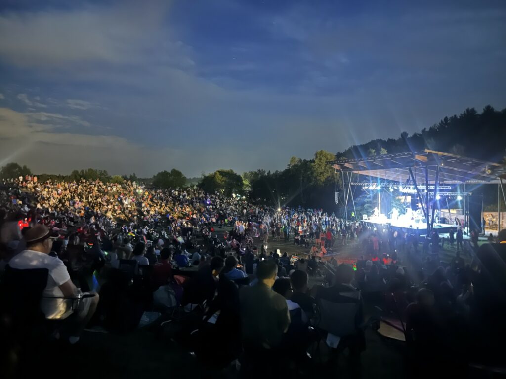 It's dusk and the bandstand is brightly lit up. We're far enough away that you can't really see the band, but some of the crowd is lit up with a spotlight.