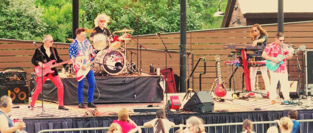 A 5-piece all male band. Right to left: bass, lead guitar and vocals, drums, keybaords, and guitar, The lead guitar player is wearing a red, white, and blue jacket with stars and stripes. Inthe very front of the photo, a few kids are hanging on a rail in front of the band listening to the music.