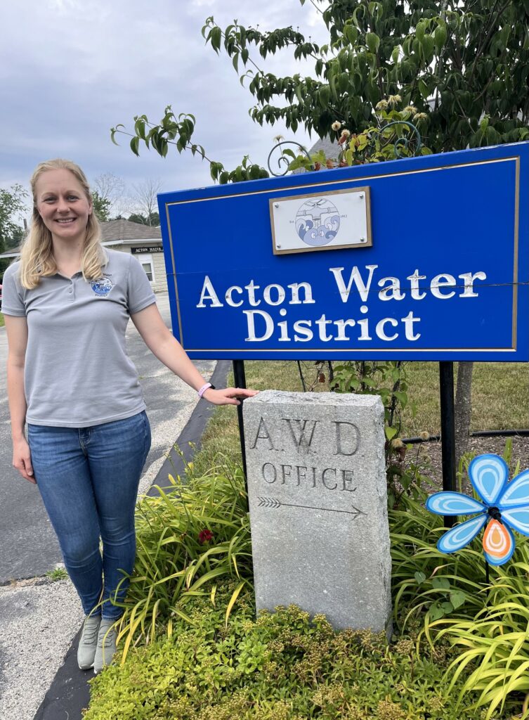 A blonde young woman stands in front of the Acton Water District sign.