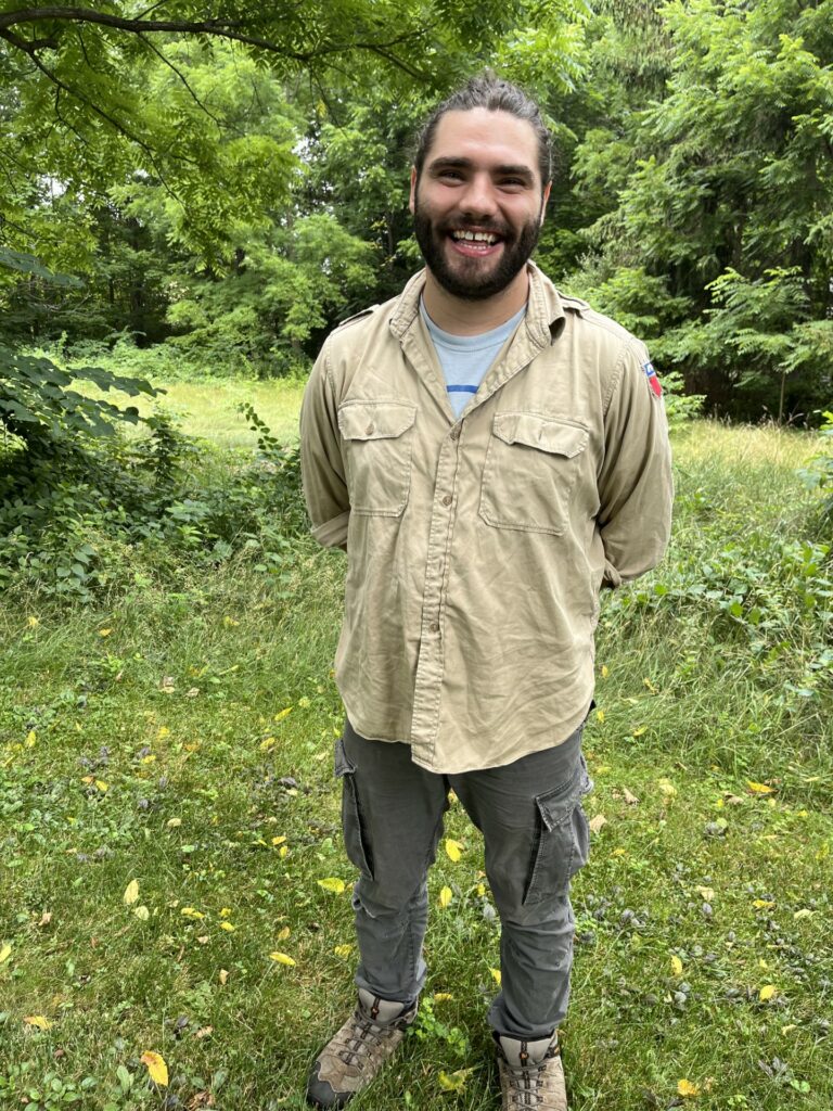 A smiling, bearded, young man stands in front of woods and a meadow.