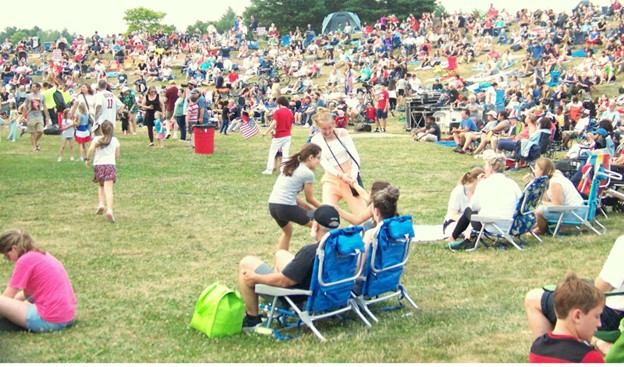 On a fields full of people with chairs and picnic blankets, two women try to tug another woman out of her chair.